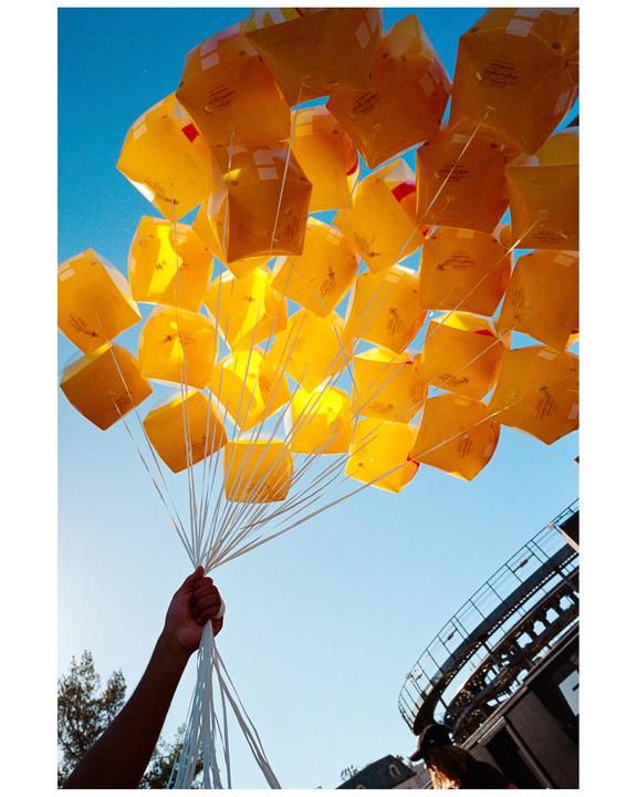 A hand holds a large bunch of yellow balloons against a clear blue sky, with a building and tree visible at the bottom.