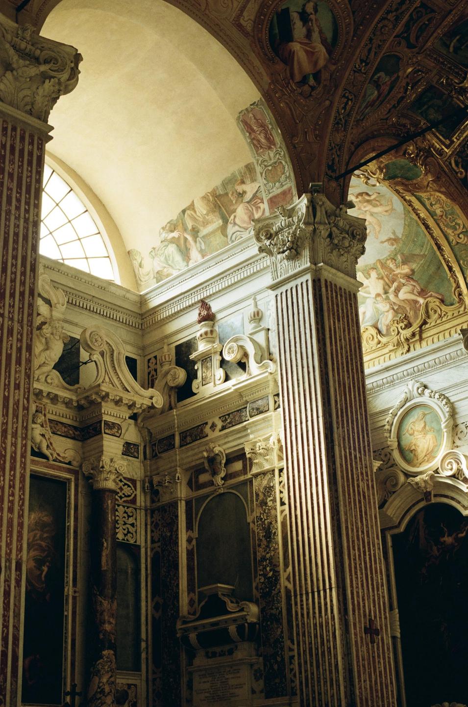 Sunlight illuminates the ornate interior of a building with painted ceilings and decorative columns.