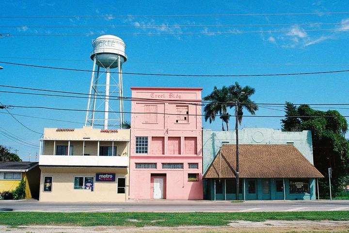 A water tower stands behind a row of colorful buildings with a bright blue sky overhead.