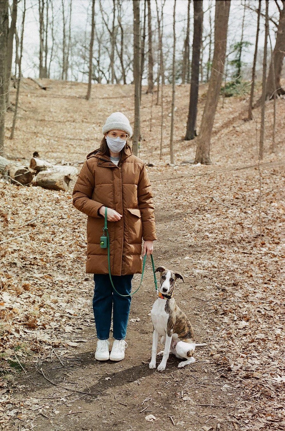 A woman in a brown coat and a dog on a leash stand on a dirt path in a forest.