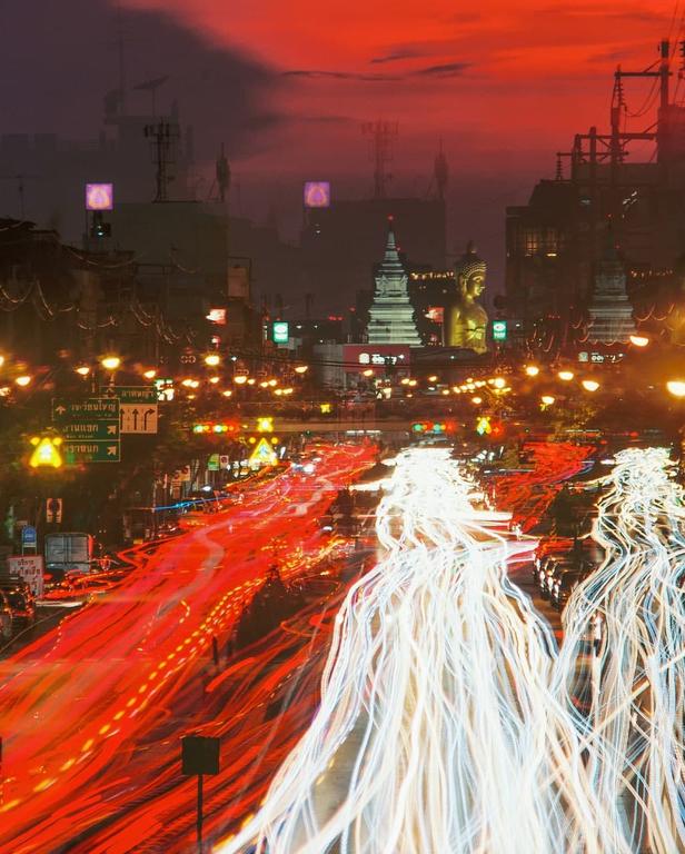 A long exposure captures light trails from vehicles on a city street at dusk, with a golden buddha statue and temple visible in the background.
