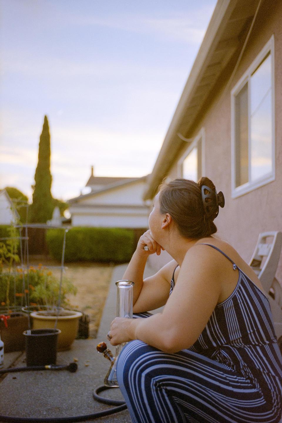 A woman in a striped jumpsuit sits on a patio with a bong, looking towards a sunset.