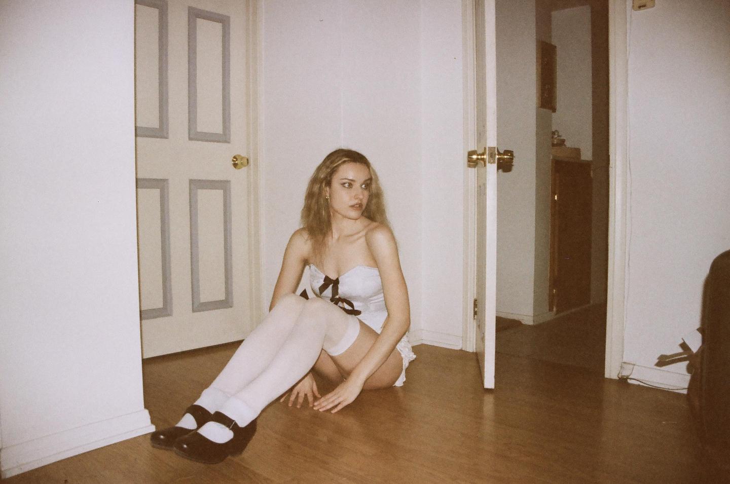 A woman in a white corset and thigh-high stockings sits on a wooden floor between two open doors.