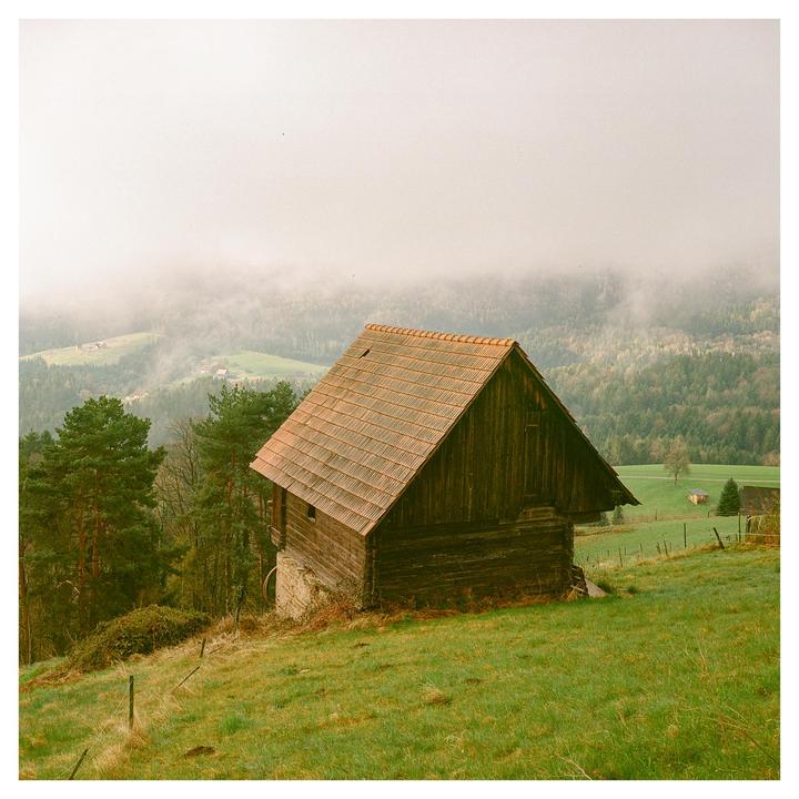 A wooden barn with a red tile roof sits on a grassy hillside overlooking a valley filled with fog and trees.