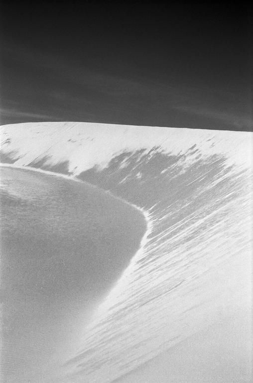 A black and white photograph captures the sweeping curves and textures of sand dunes under a dark sky.