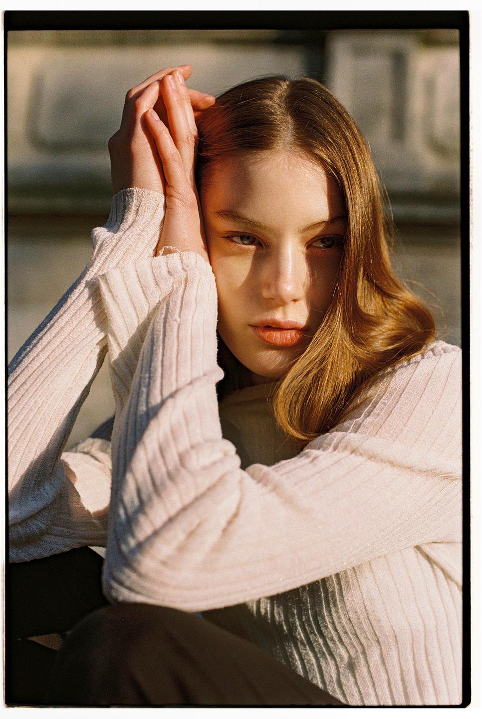 A young woman with long hair and a light-colored sweater poses with her hands clasped above her head.