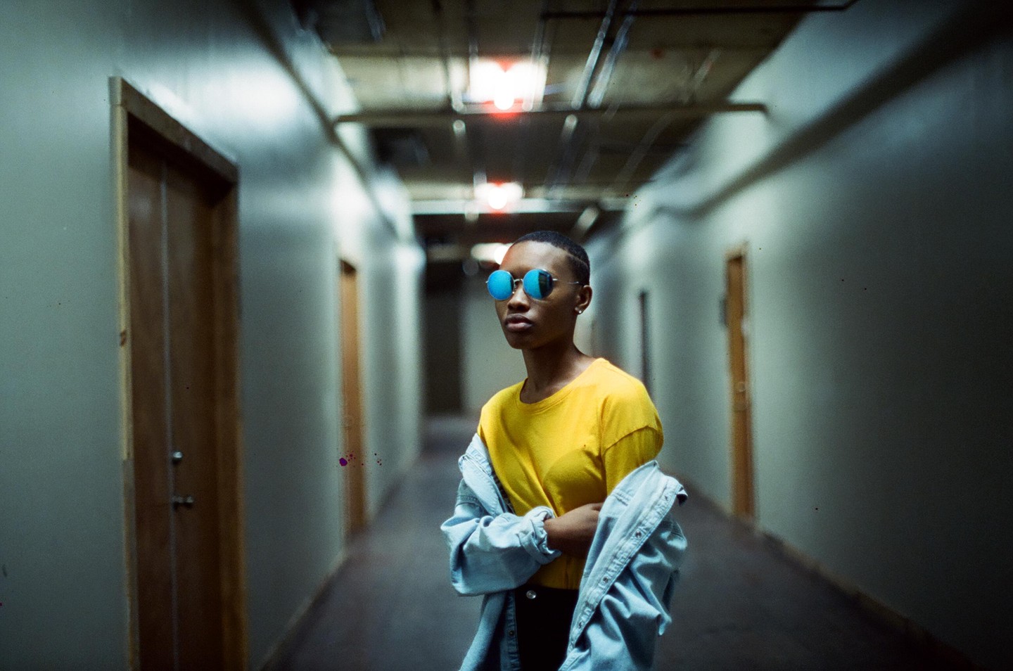 A woman in a yellow shirt and blue sunglasses stands in a long hallway with doors on either side.
