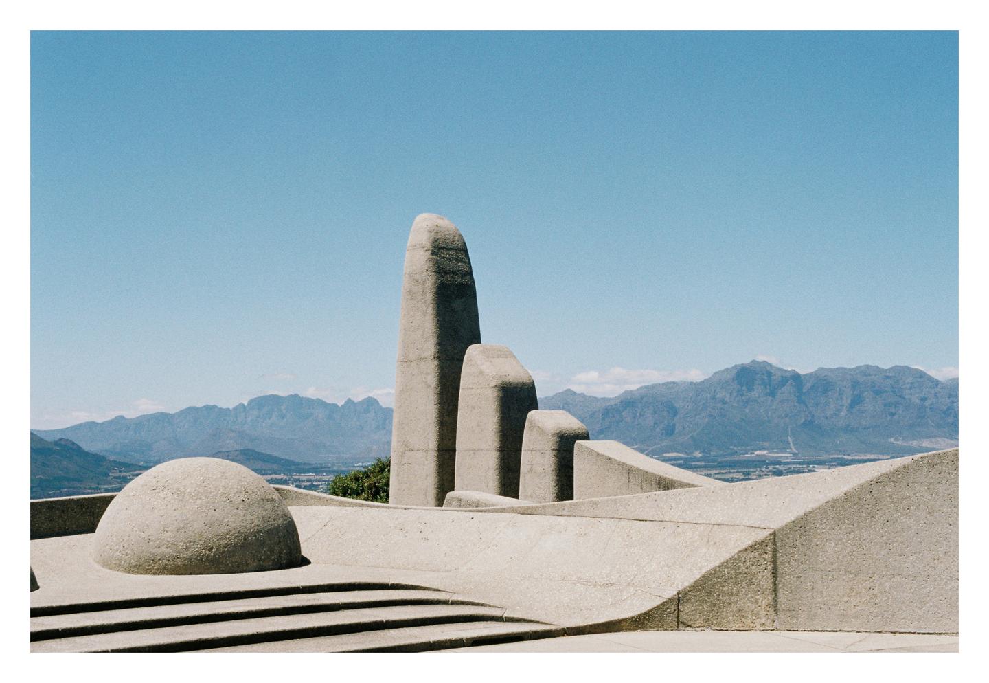 A concrete monument with a rounded dome and tall, angular structures stands before a mountain range under a clear sky.