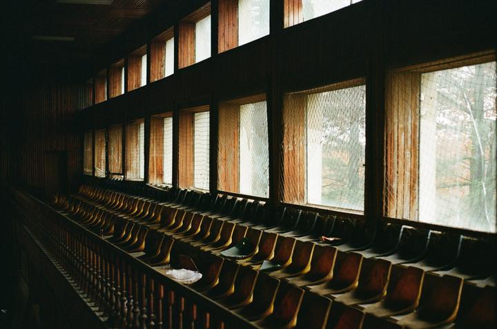 Rows of empty wooden seats face a wall of windows with broken panes in an abandoned sports complex.
