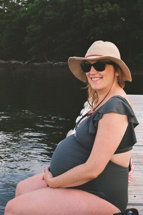 A pregnant woman wearing a sun hat and sunglasses sits on a wooden dock next to water with trees in the background.