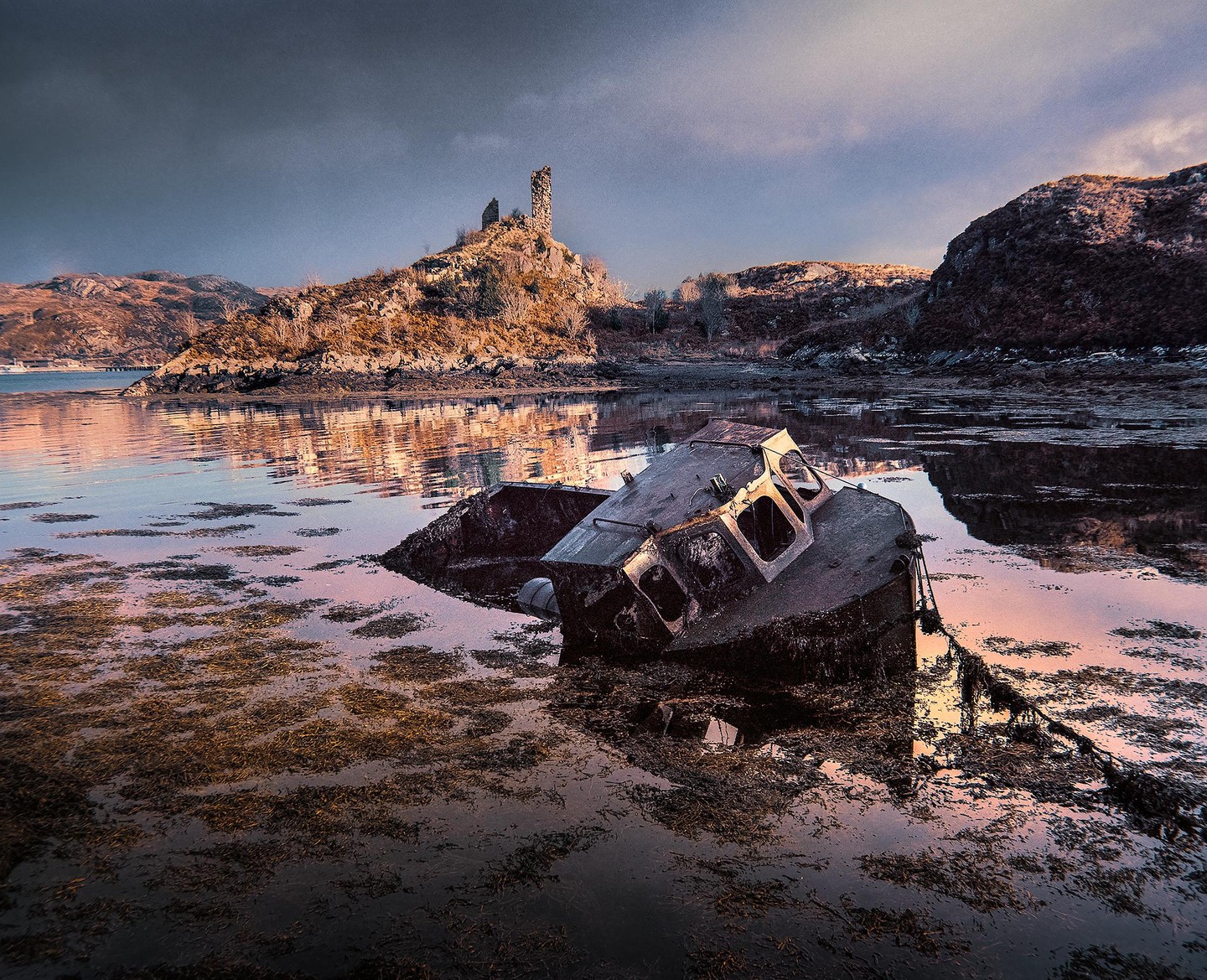 A derelict boat lies half-submerged in shallow water with a ruined castle on a hill in the background.