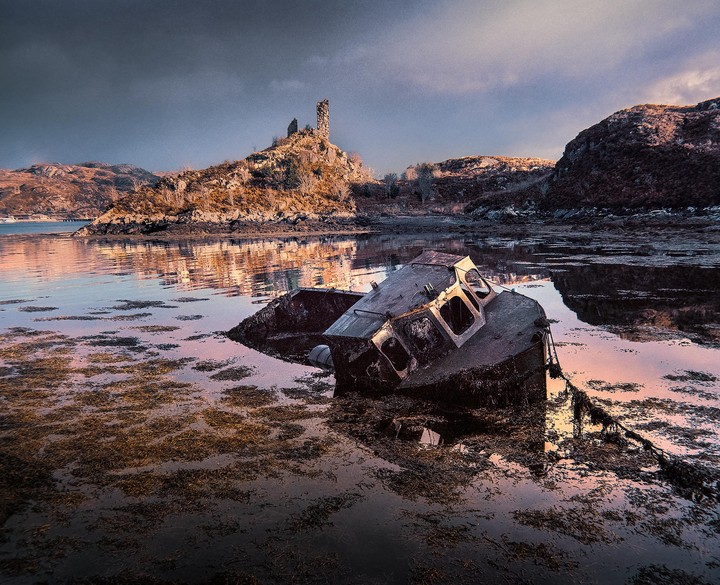 A derelict boat lies half-submerged in shallow water with a ruined castle on a hill in the background.