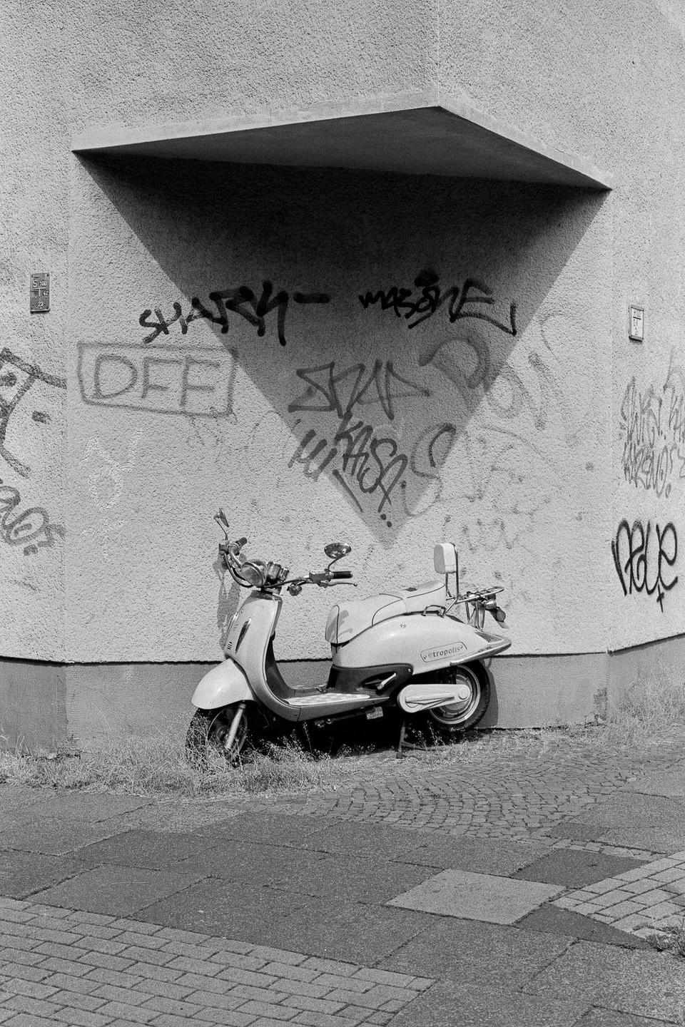A scooter is parked on a sidewalk next to a graffiti-covered wall with a triangular shadow.
