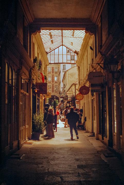 People walk through a narrow, sunlit alley with shops on both sides and a glass roof overhead.