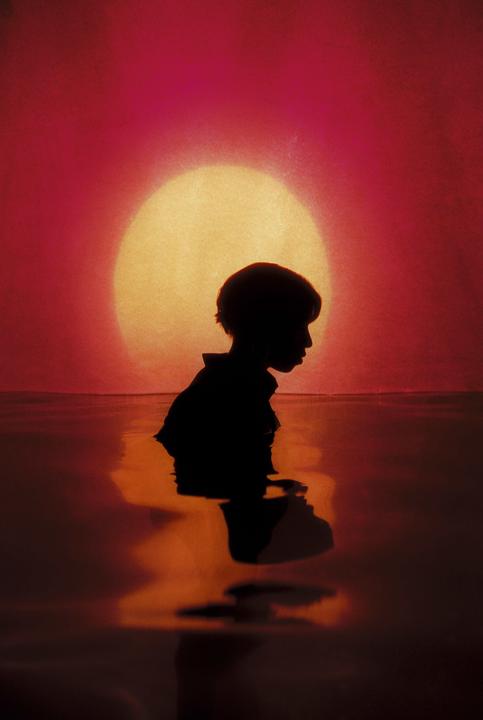 A child is silhouetted against a large, bright sun as they stand in water.