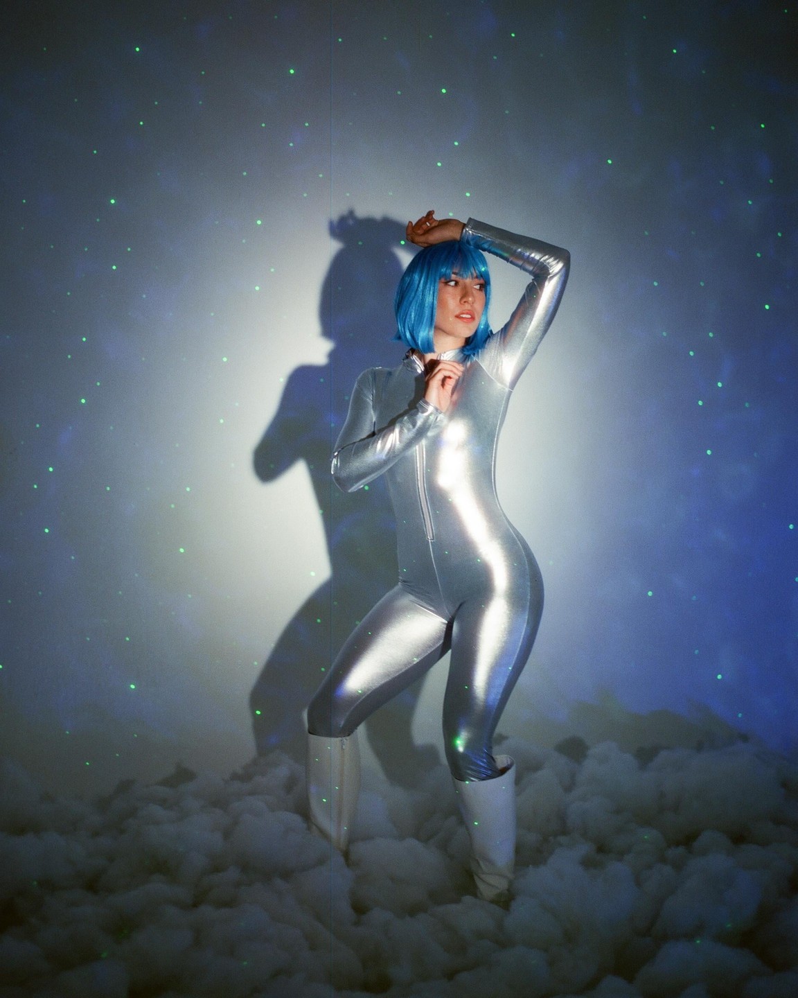 A woman in a silver jumpsuit and blue wig stands in a cloud-filled room with green lights projected on the wall.
