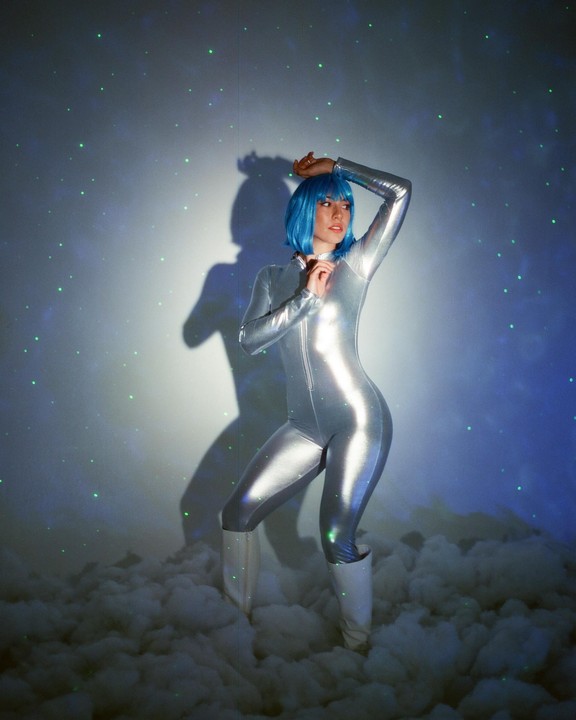 A woman in a silver jumpsuit and blue wig stands in a cloud-filled room with green lights projected on the wall.