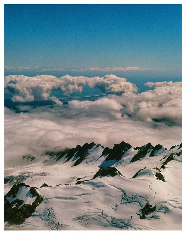 Snow-covered mountain peaks are visible below a layer of clouds, with a body of water and more clouds in the distance under a clear blue sky.