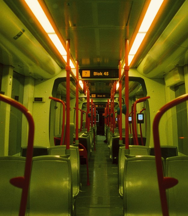 The interior of an empty train car is illuminated by orange lights, with rows of seats and red poles extending down the aisle.