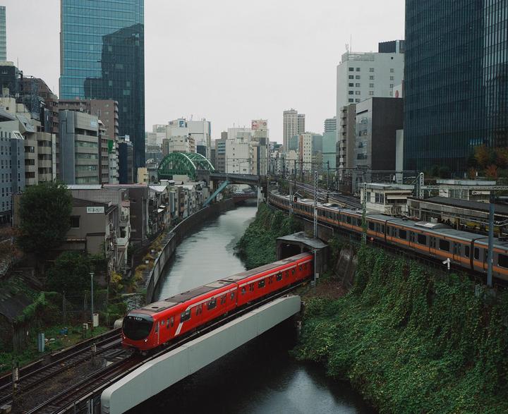 A red train crosses a bridge over a river, with another train on tracks above and buildings in the background.