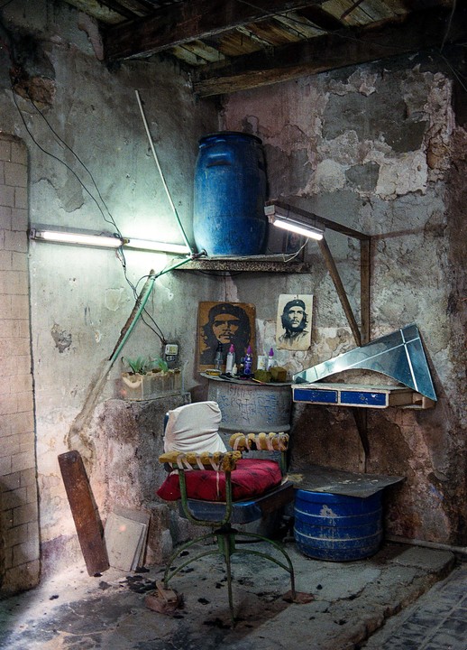 A barber chair with red cushions sits in a small, cluttered room with two portraits of Che Guevara on the wall.