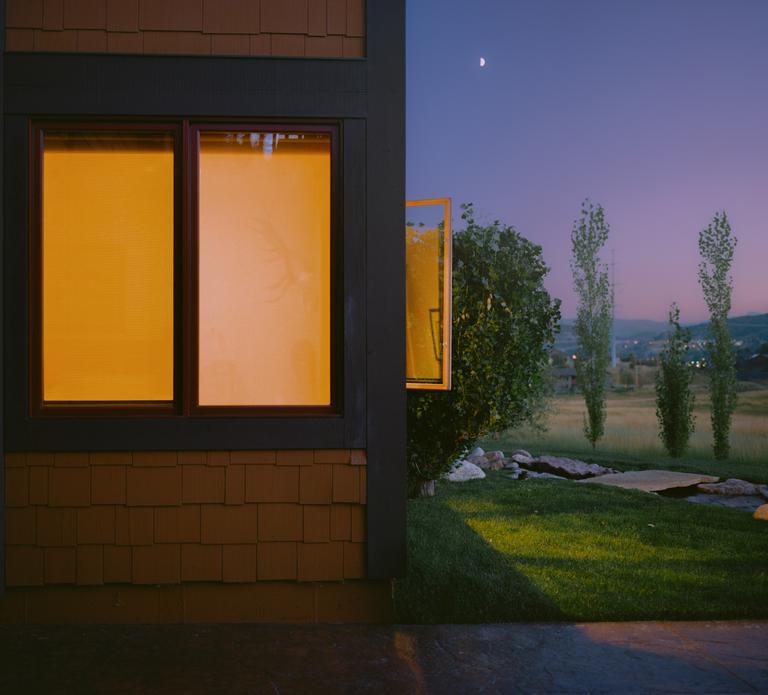 A house with illuminated windows stands next to a grassy field under a twilight sky with a crescent moon.