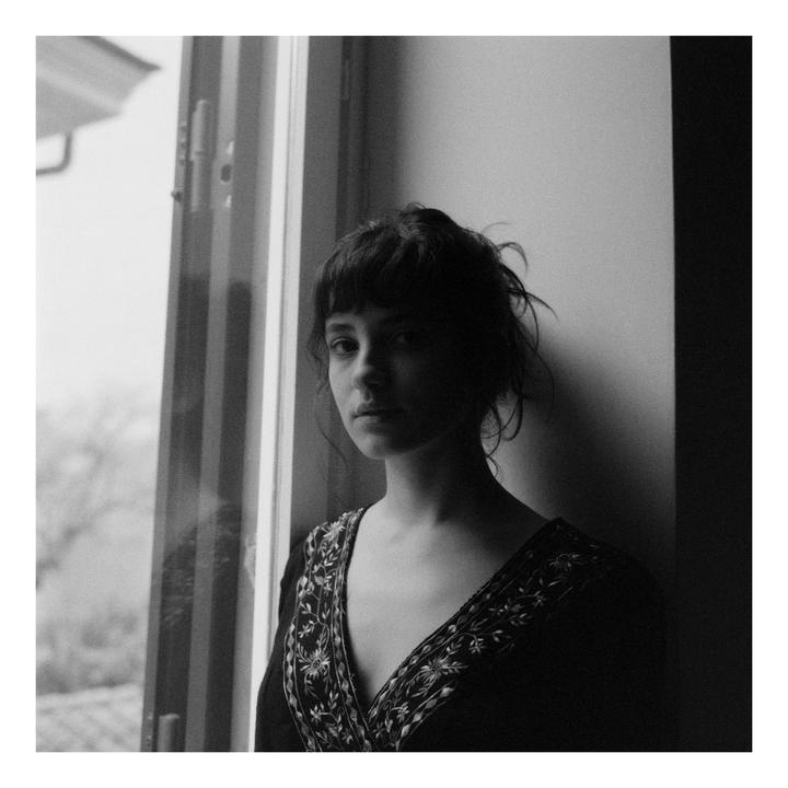 A woman with bangs and messy hair stands next to a window, looking directly at the viewer.