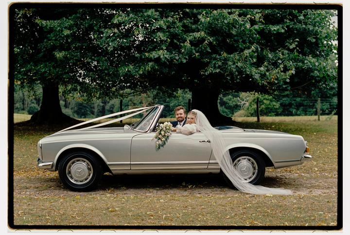 A couple sits in a silver convertible car decorated with white ribbons and a bouquet, parked under a large tree.