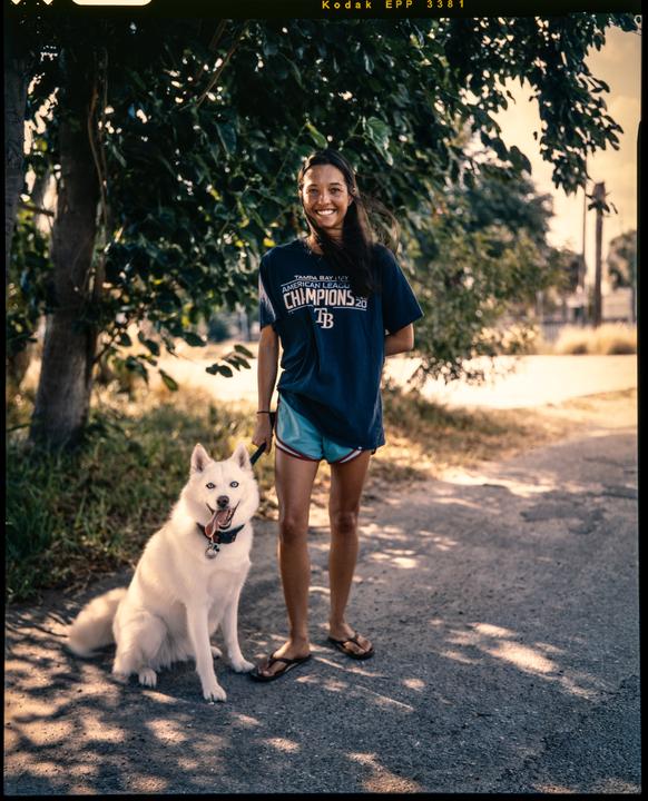 A woman and a white dog stand on a paved path under dappled sunlight.