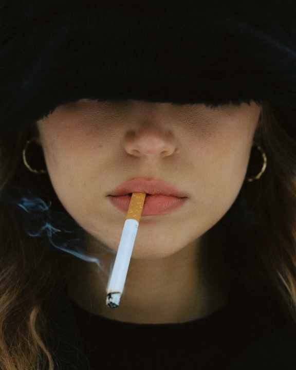 A woman with a black hat covering her eyes holds a lit cigarette in her mouth, with smoke curling upwards.