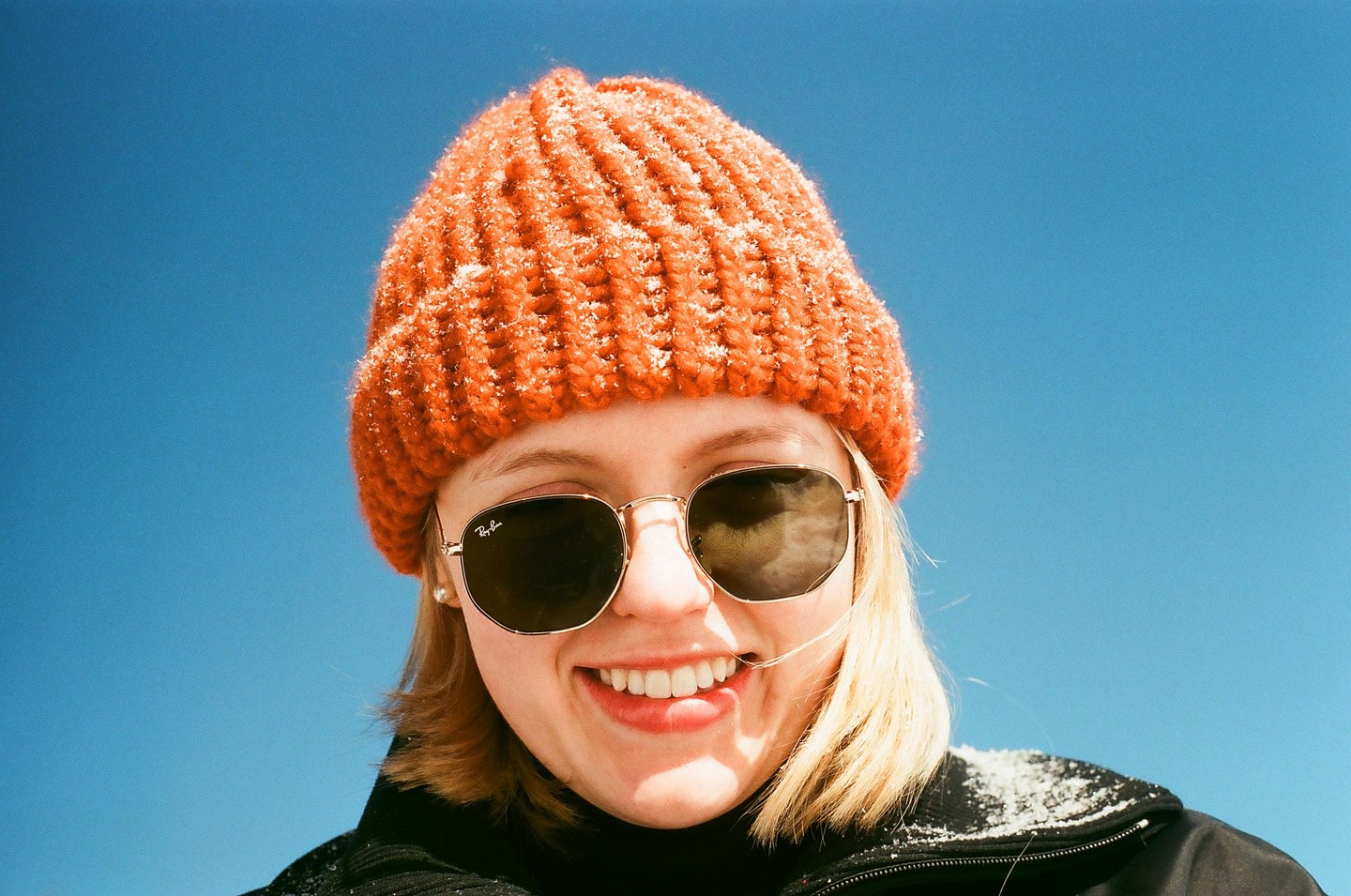 A woman wearing an orange beanie and sunglasses smiles at the camera against a clear blue sky.