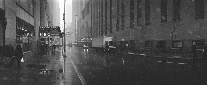 A panoramic street scene shows a city street with buildings, cars, and pedestrians during a snowfall.