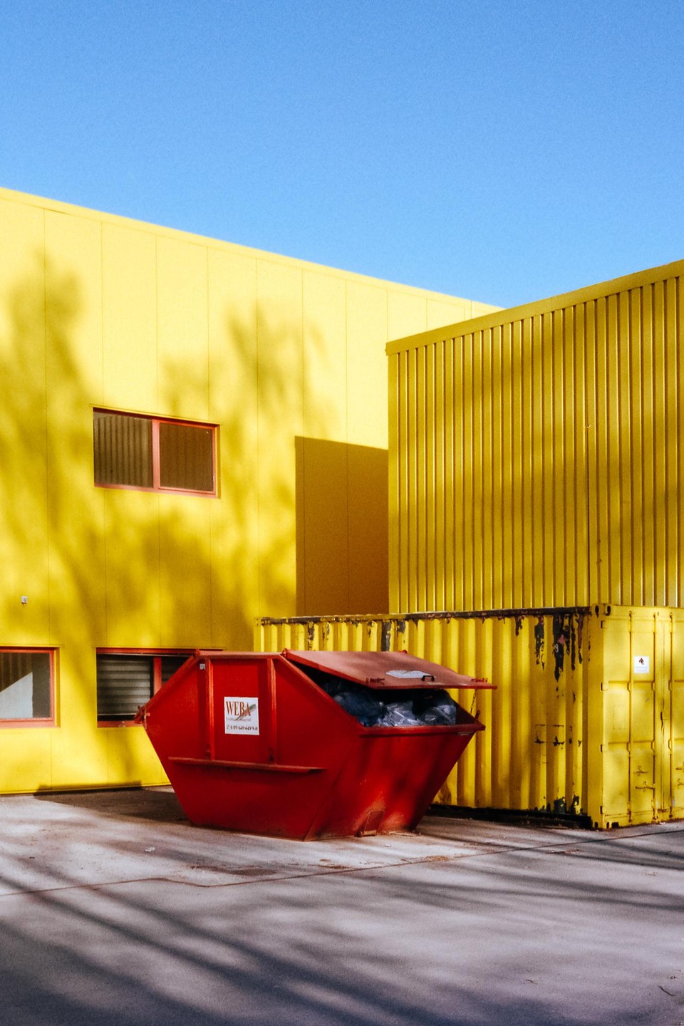 A red dumpster sits in front of a bright yellow building with shadows cast across its facade.