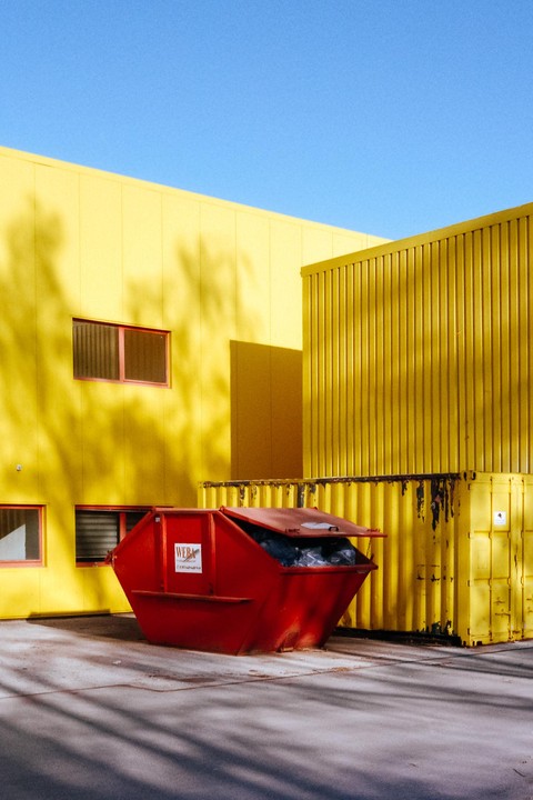 A red dumpster sits in front of a bright yellow building with shadows cast across its facade.