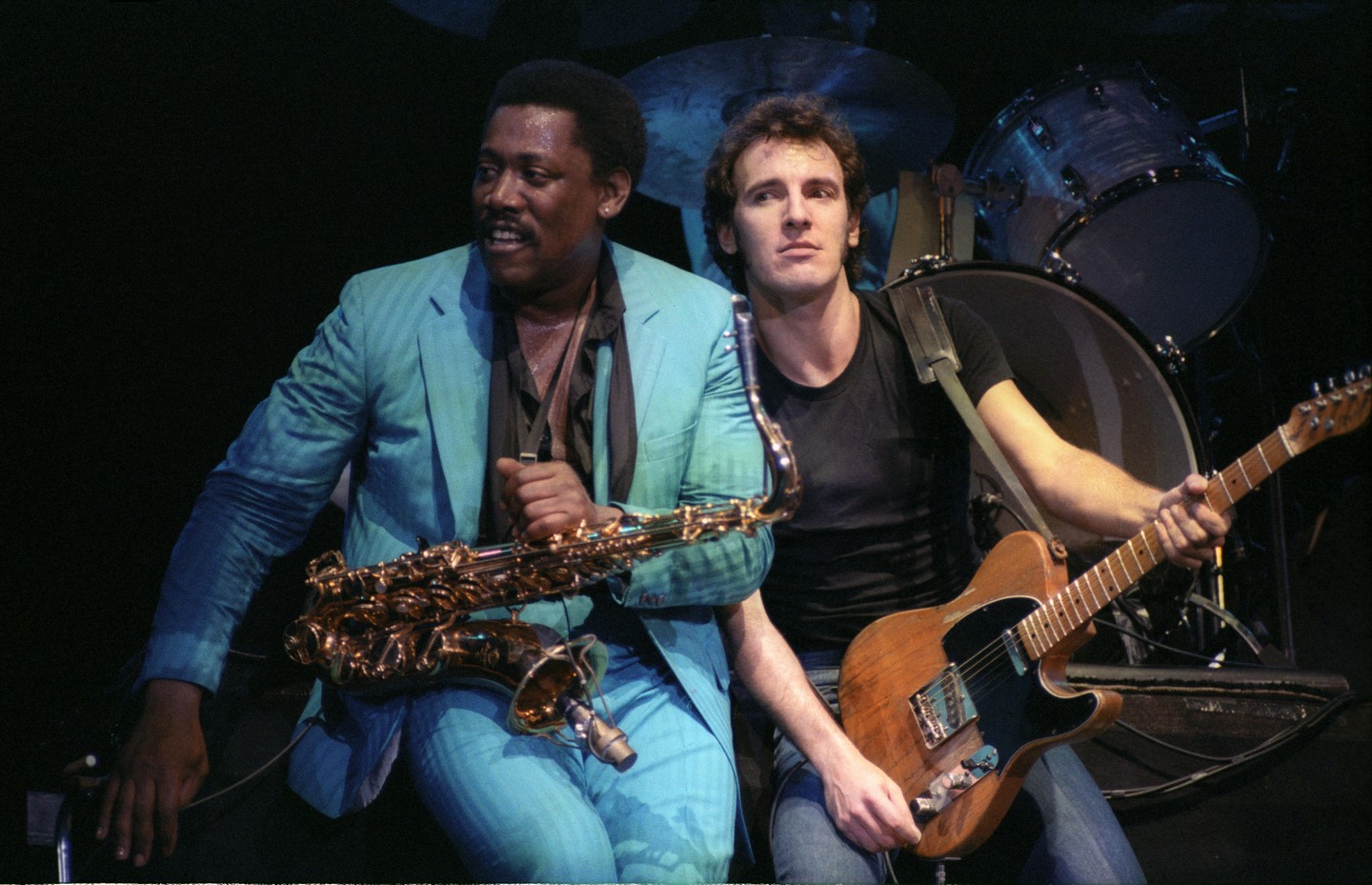 Two men are performing on stage, one holding a saxophone and the other a guitar.