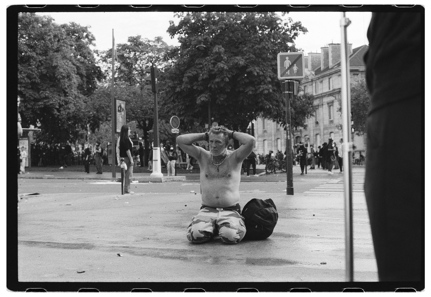 A shirtless man kneels on a wet street with his hands behind his head, next to a backpack, with a crowd of people and trees in the background.