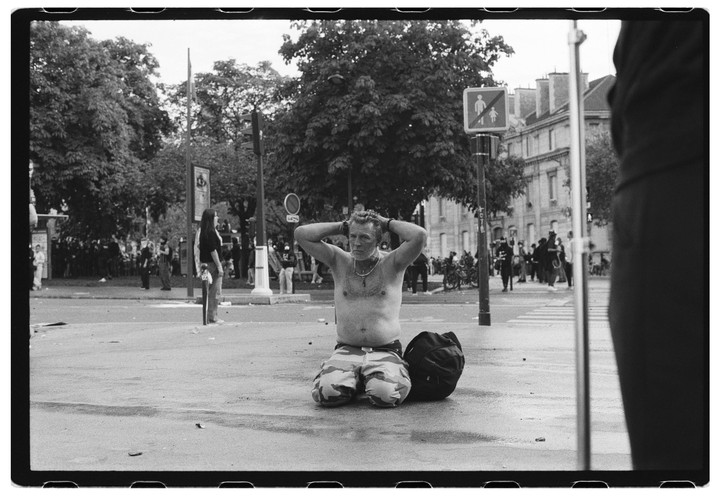 A shirtless man kneels on a wet street with his hands behind his head, next to a backpack, with a crowd of people and trees in the background.