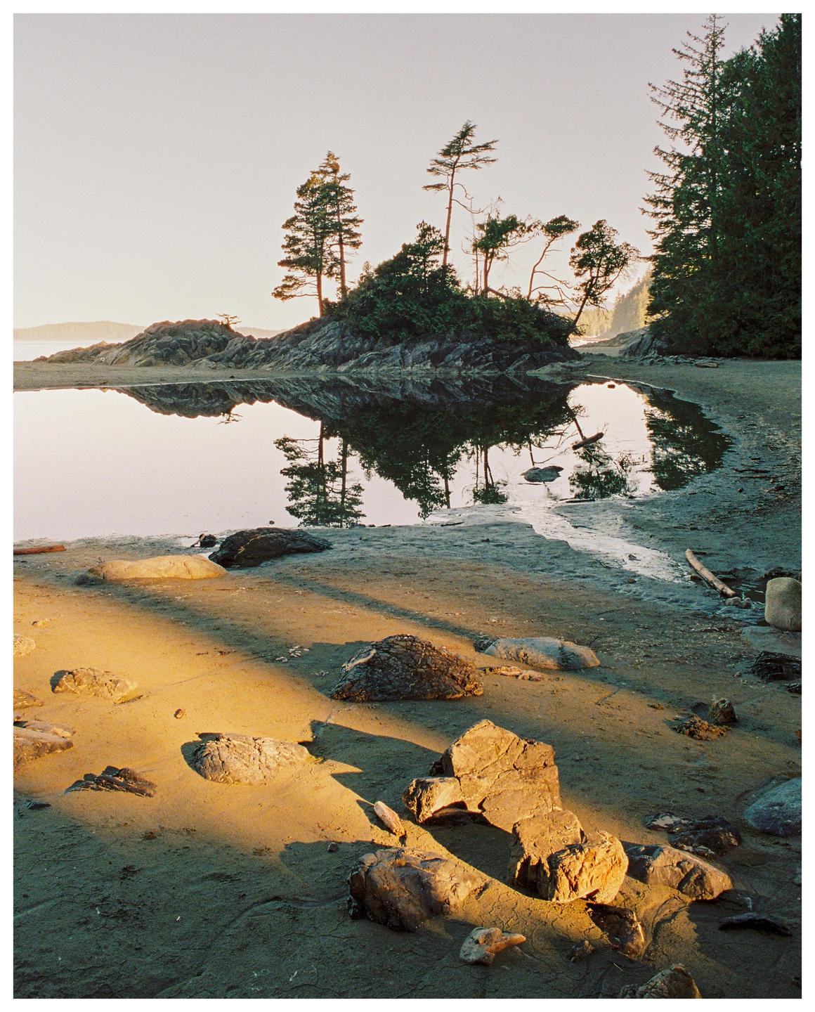 A sandy beach with rocks and a tidal pool reflecting trees and a rocky island under a bright sky.