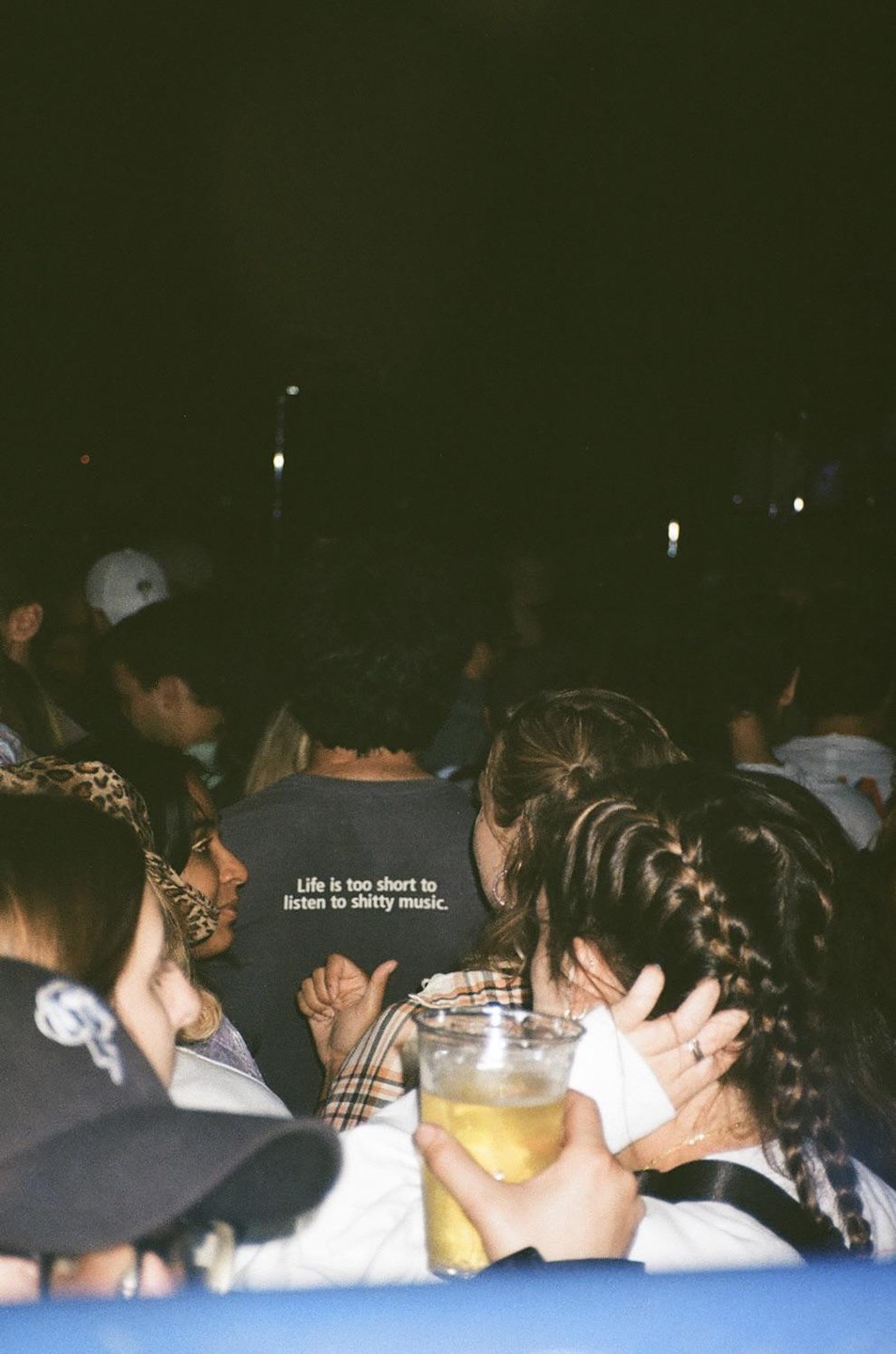 A person wearing a shirt that says "Life is too short to listen to shitty music" is visible in a crowd at night.