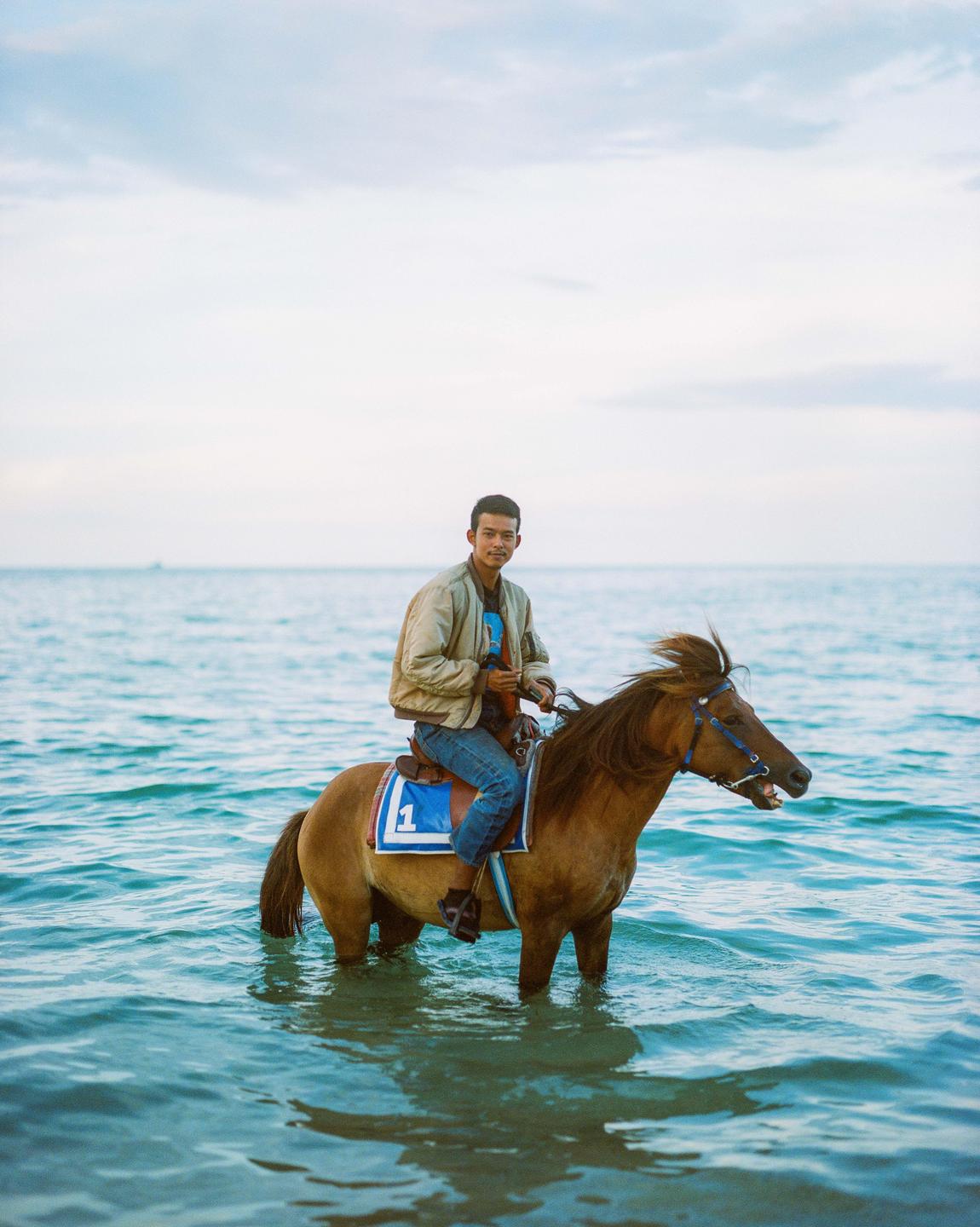 A man rides a horse in the ocean with a cloudy sky in the background.