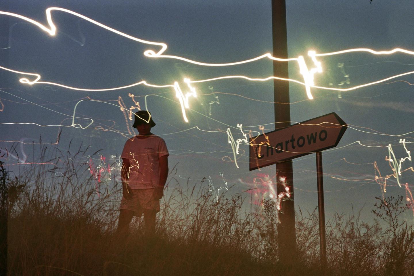 A man stands in tall grass next to a Chartowo sign with light trails streaking across the night sky.
