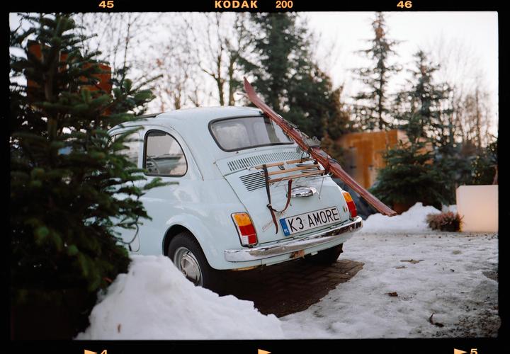 A light blue car with skis on its back is parked in a snowy area next to a pine tree.