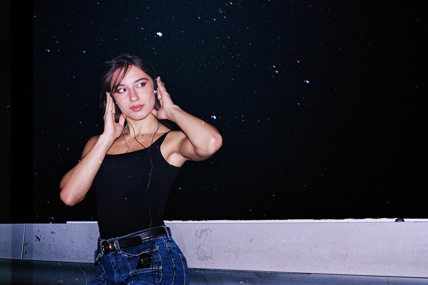A woman wearing headphones looks to the side against a starry night sky.