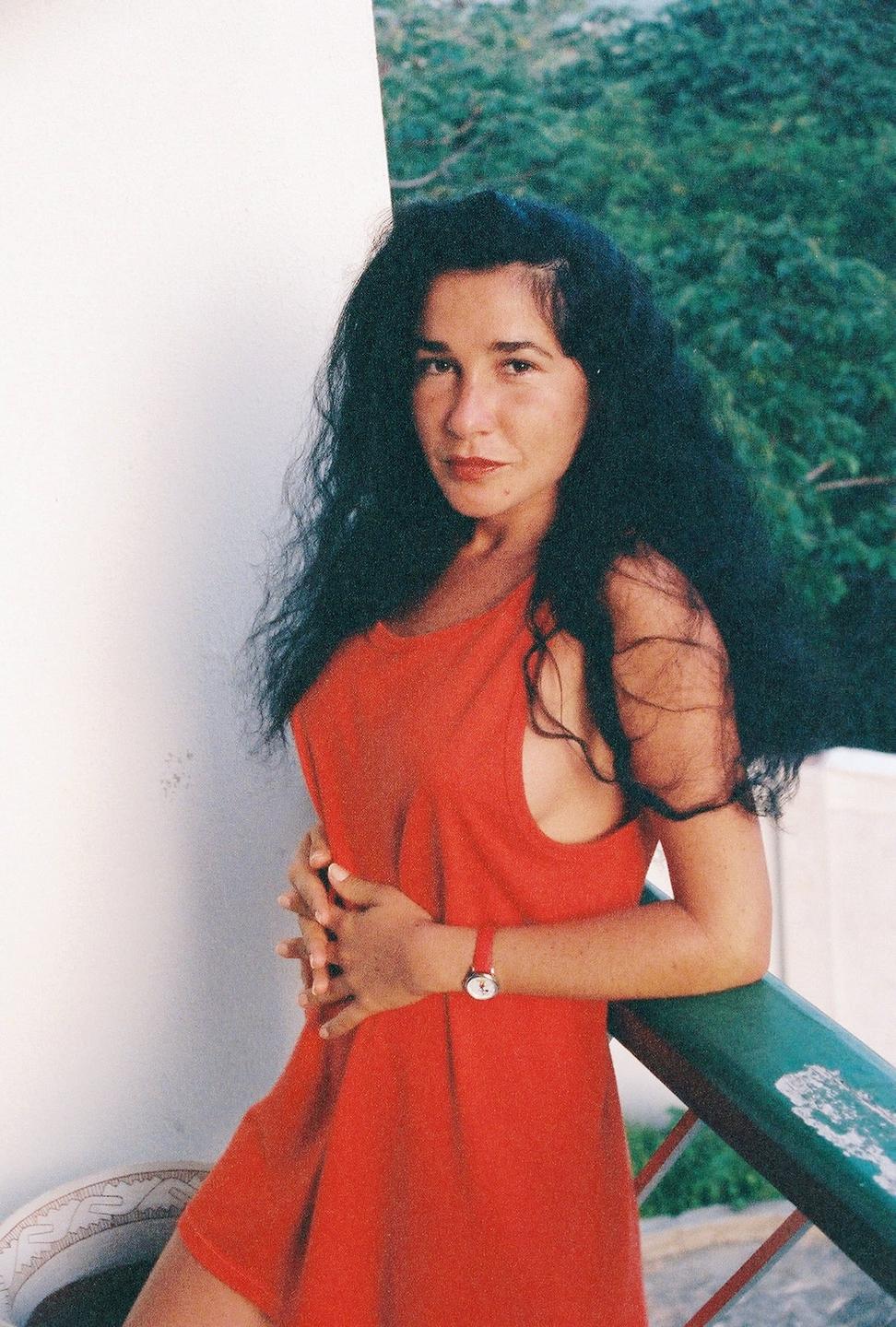 A woman with long dark hair and a red dress leans against a railing with green foliage in the background.