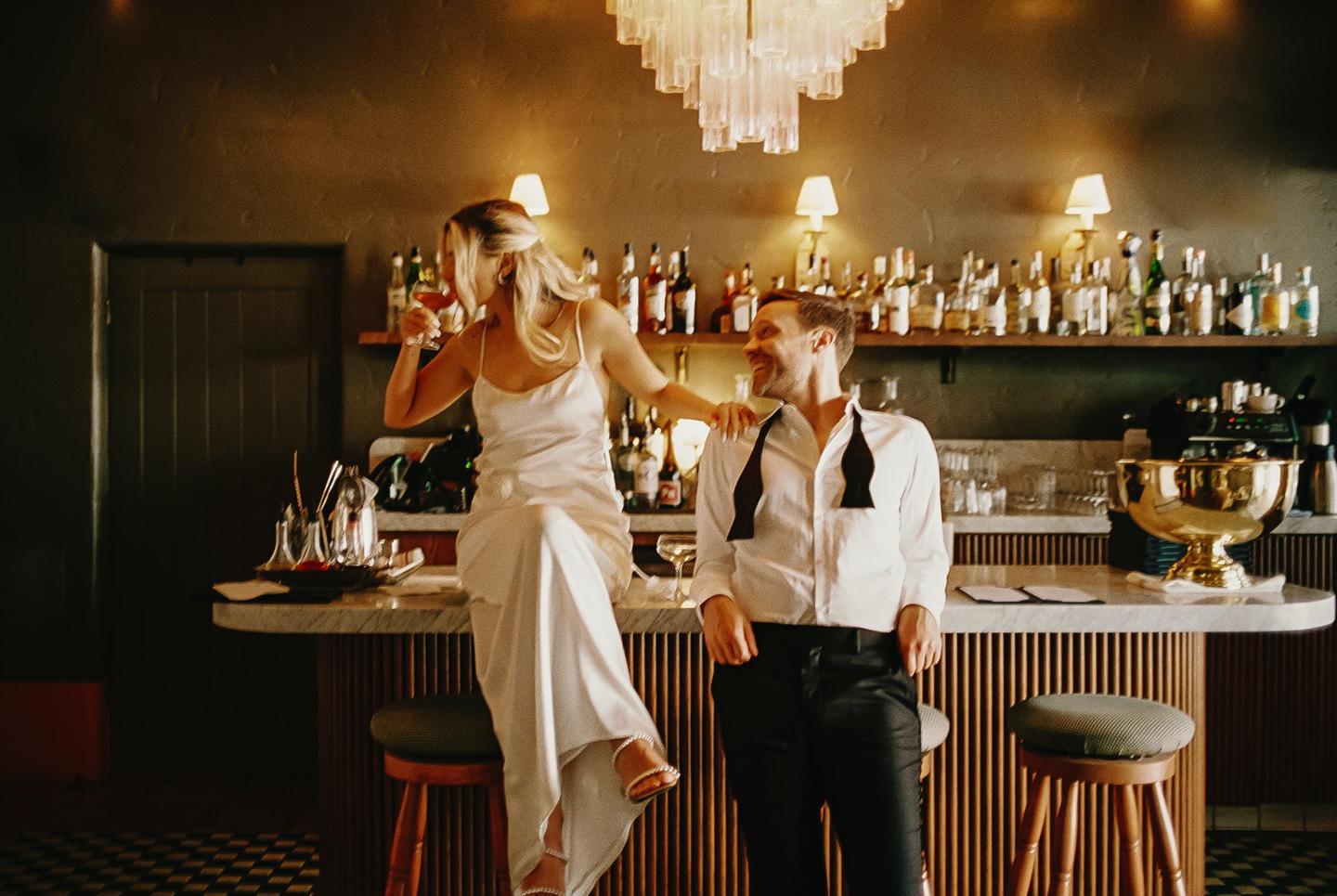 A woman in a white dress sits on a bar while a man in a white shirt and black pants stands next to her.