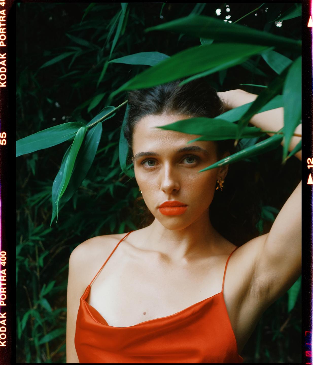 A woman in a red dress with red lipstick poses among green foliage.