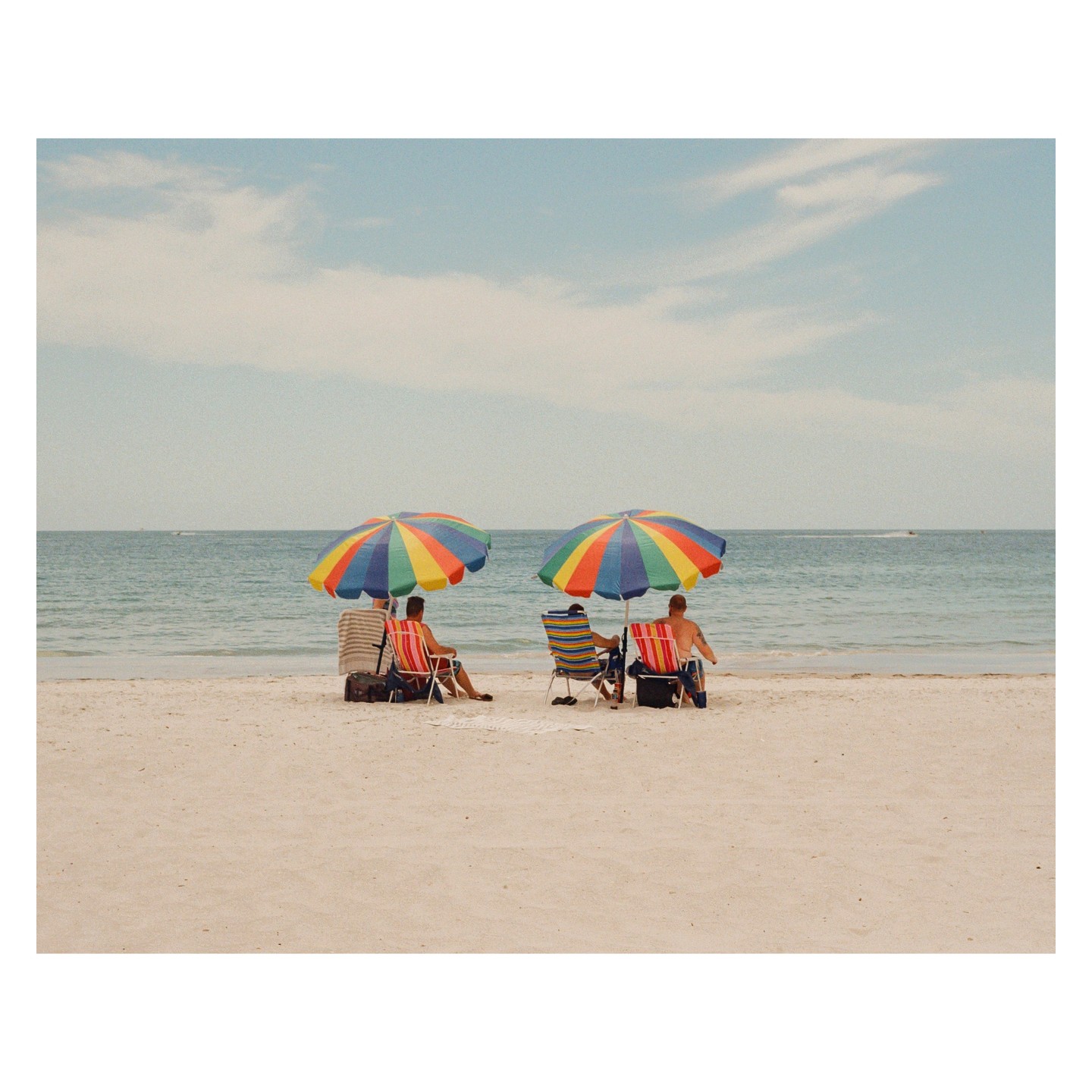 Two men sit in beach chairs under colorful umbrellas on a sandy beach facing the ocean.