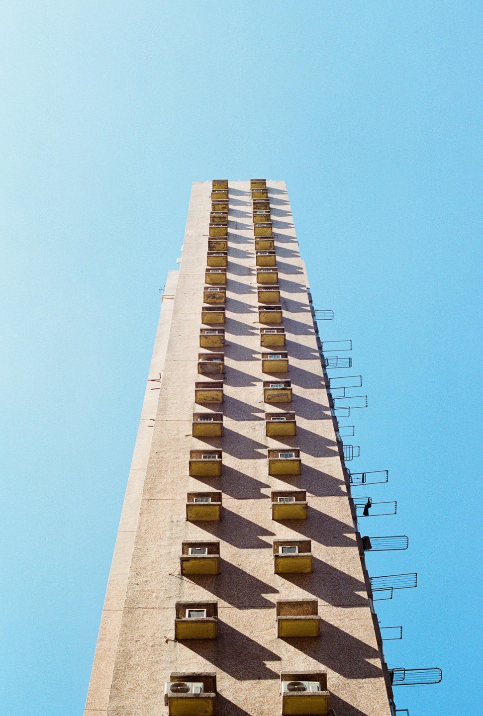 A tall, narrow building with many small balconies and air conditioning units is shown against a clear blue sky.