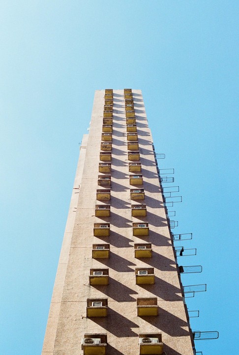 A tall, narrow building with many small balconies and air conditioning units is shown against a clear blue sky.