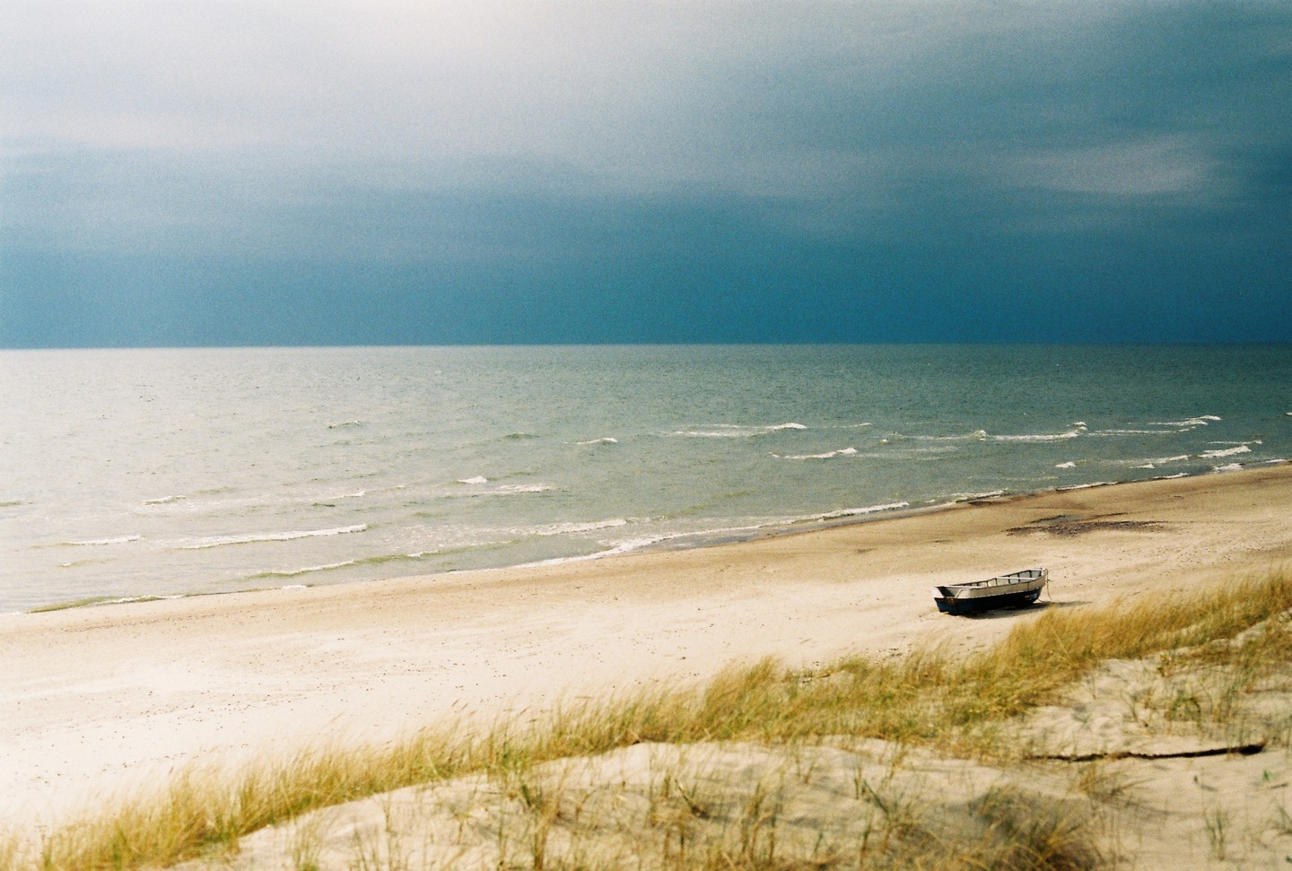 A small boat rests on a sandy beach with gentle waves under a dramatic, dark blue sky.